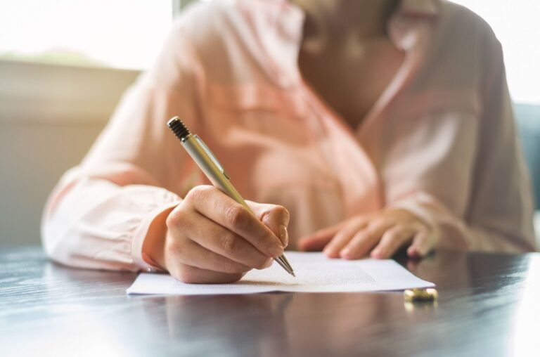 Imagem de uma mulher, sem aparecer o rosto, sentada e assinando um documento.