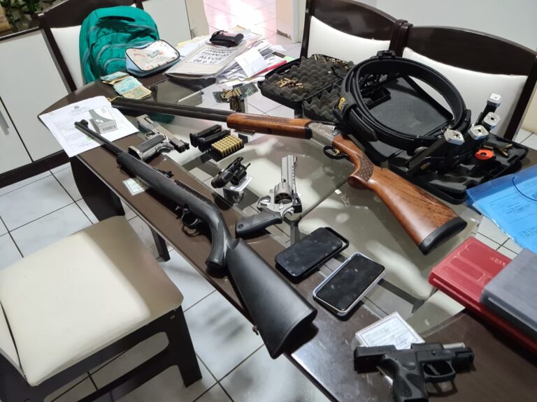 Imagem de várias armas, munições, documentos, aparelhos celulares e dinheiro sobre uma mesa de vidro.