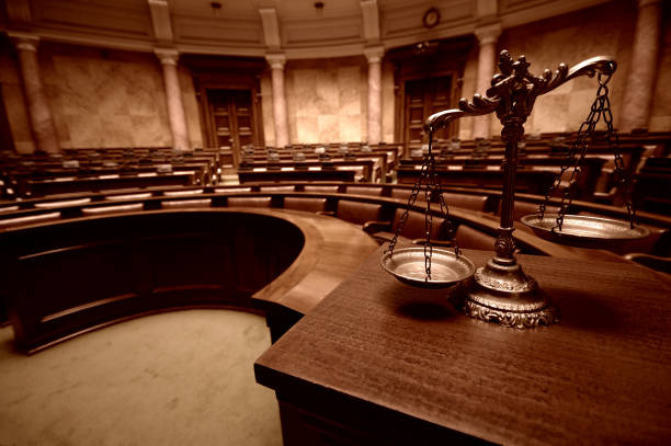 Imagem de uma sala de júri, vazia e, em primeiro plano, em cima de uma mesa em madeira, uma balança representando a Justiça.