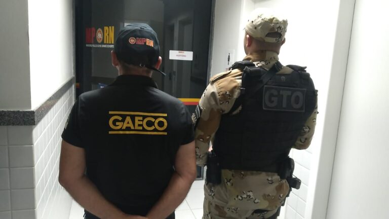 Imagem de um agente do Gaeco e de um policial do GTO, ambos de costas, em pé, em frente a uma porta de vidro.
