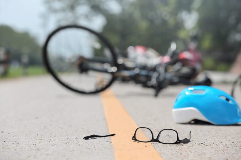 Imagem de bicicleta deitada em uma via de trânsito com capacete, óculos e itens pessoais espalhados após o acidente.