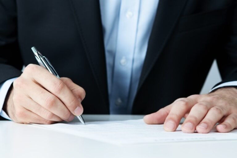 Foto mostra apenas o peitoral de um homem vestido com um terno enquanto apoia uma mão na mesa e a outra mão assina um documento