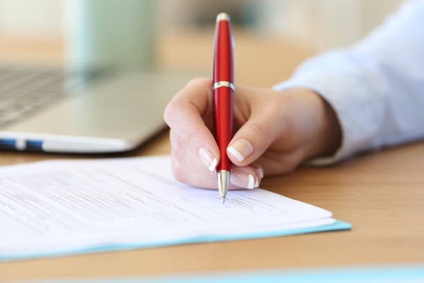 Imagem de uma mão feminina assinando um documento em cima de uma mesa.