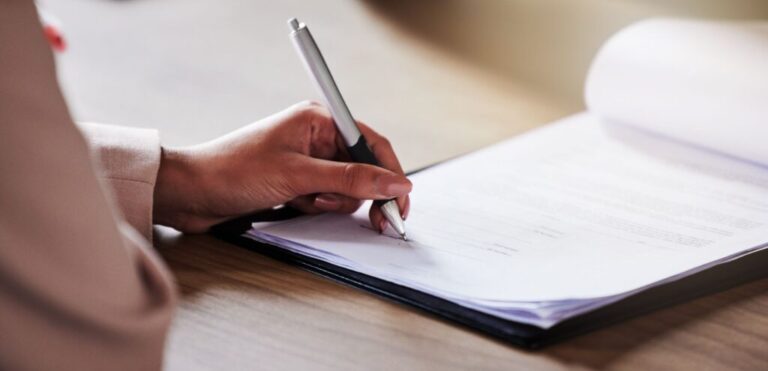 Imagem da mão de uma mulher, segurando uma caneta e assinando um papel.