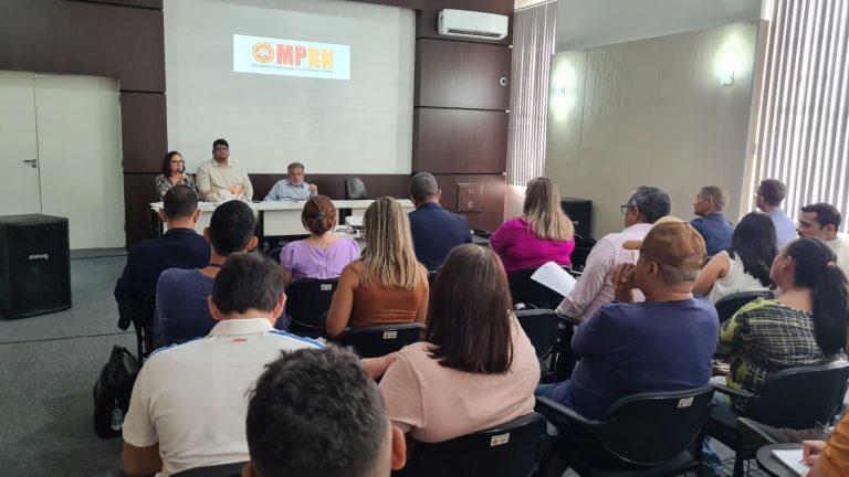 Foto de um auditório, com pessoas de costas,sentadas, em primeiro plano e ao fundo os promotores de Justiça sentados em uma meda, de frente para a plateia