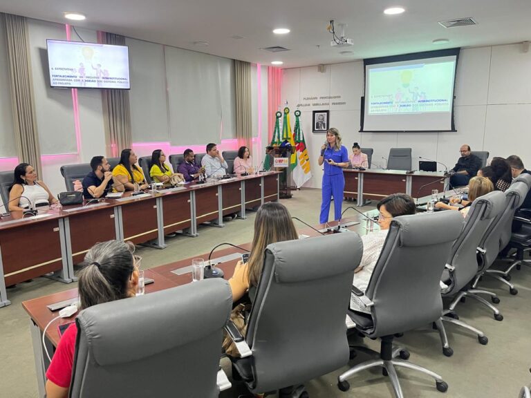 Imagem mostra um plenario com pessoas sentadas às mesas e uma mulher branca, vestida de azul, com um microfone, falando para os presente