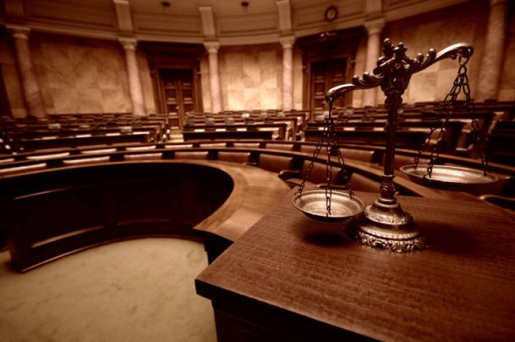 Imagem de uma sala de júri, vazia e, em primeiro plano, em cima de uma mesa em madeira, uma balança representando a Justiça.