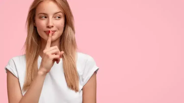 Indoor shot of beautiful female looks mysteriously aside, has intriguing look, asks to be quiet, dressed in casual clothes, stands against pink background with blank copy space for your advertisement