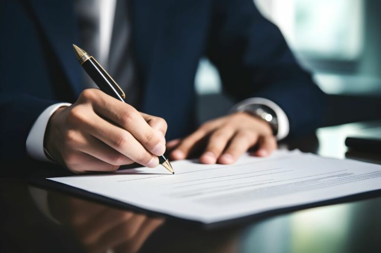 Imagem de um homem, vestido de terno e gravata, sentado, assinando um documento que está sobre uma mesa.