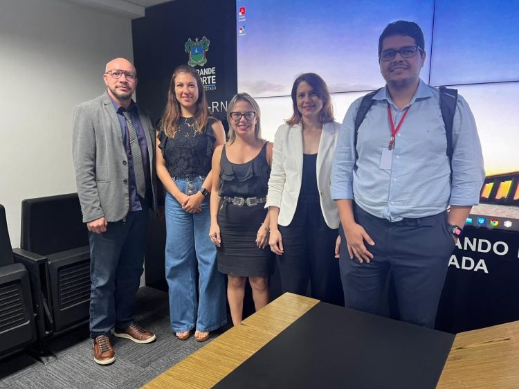 Imagem de cinco pessoas, sendo dois homens e três mulheres, todos em pé, em uma sala, posando para a foto.