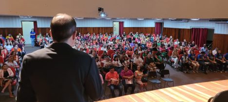 Imagem de um auditório repleto de pessoas e um homem, no palco, falando para essa plateia.