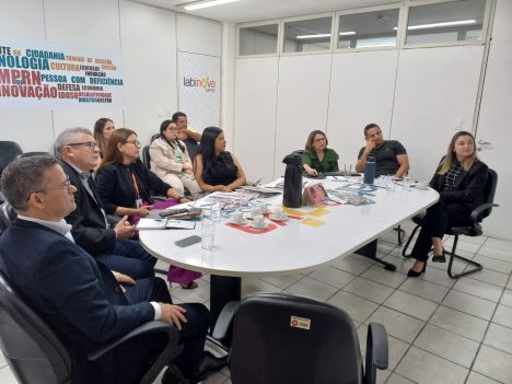 Imagem com várias pessoas sentadas ao redor de uma mesa oval, em uma sala, participando de uma reunião.