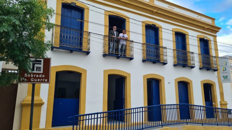 Imagem que mostas um sobrado de dois andares pintado de branco, com portas azuis e molduras das portas amarelas, tem dois homnes em uma das sacadas no andar superior.