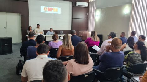 Foto de um auditório, com pessoas de costas,sentadas, em primeiro plano e ao fundo os promotores de Justiça sentados em uma meda, de frente para a plateia