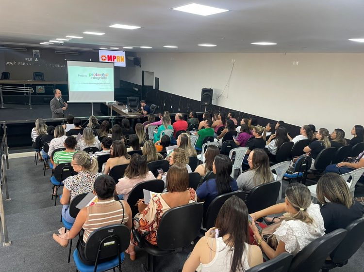 Imagem mostra um auditório da perspectiva de quem está atrás de todas as fileiras de cadeiras, todas ocupadas por pessoas. Ao fundo, perto do palco tem um banner com o slogan do MPRN e um homem branco em pé falando para o público.