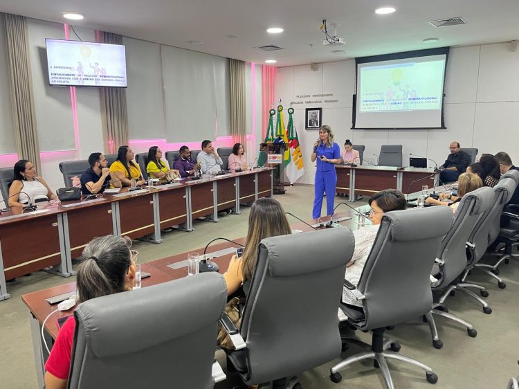 Imagem mostra um plenario com pessoas sentadas às mesas e uma mulher branca, vestida de azul, com um microfone, falando para os presente